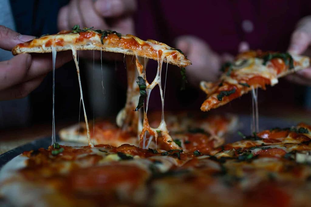 Three people are holding slices of pizza with melted cheese stretching from the pizza, topped with basil and pepperoni at Caliente Pizza & Drafthouse, a popular craft beer bar in Pittsburgh.