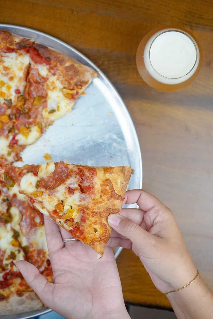 A person holds a slice of pizza over a pizza tray on a wooden table. A glass of beer is nearby.