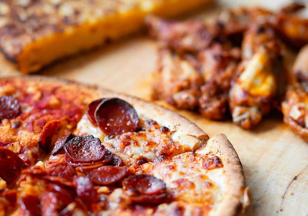 Close-up of a pepperoni pizza with a crispy crust from Pittsburgh Pizzeria, a stack of chicken wings, and a breaded item in the background on a wooden surface.