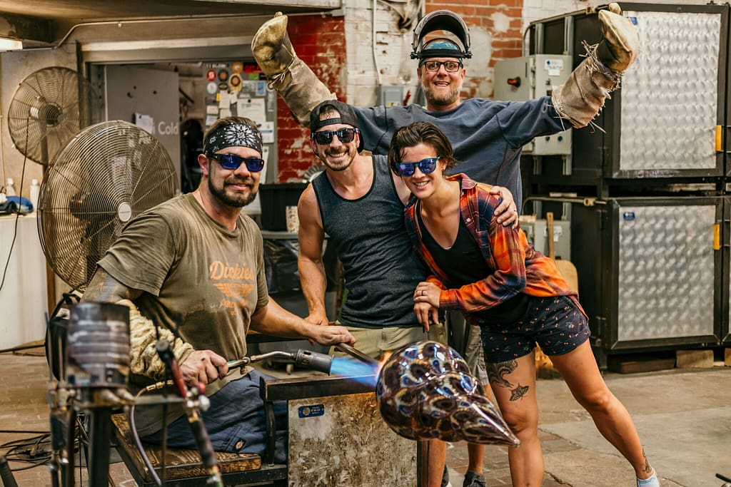 Dan Alexander sitting down in the hot glass studio, torching a large glass work, surrounded by two friends, with Scott Krenitsky, hot glass studio manager raising his arms in victory in the background. All are smiling for the photo.