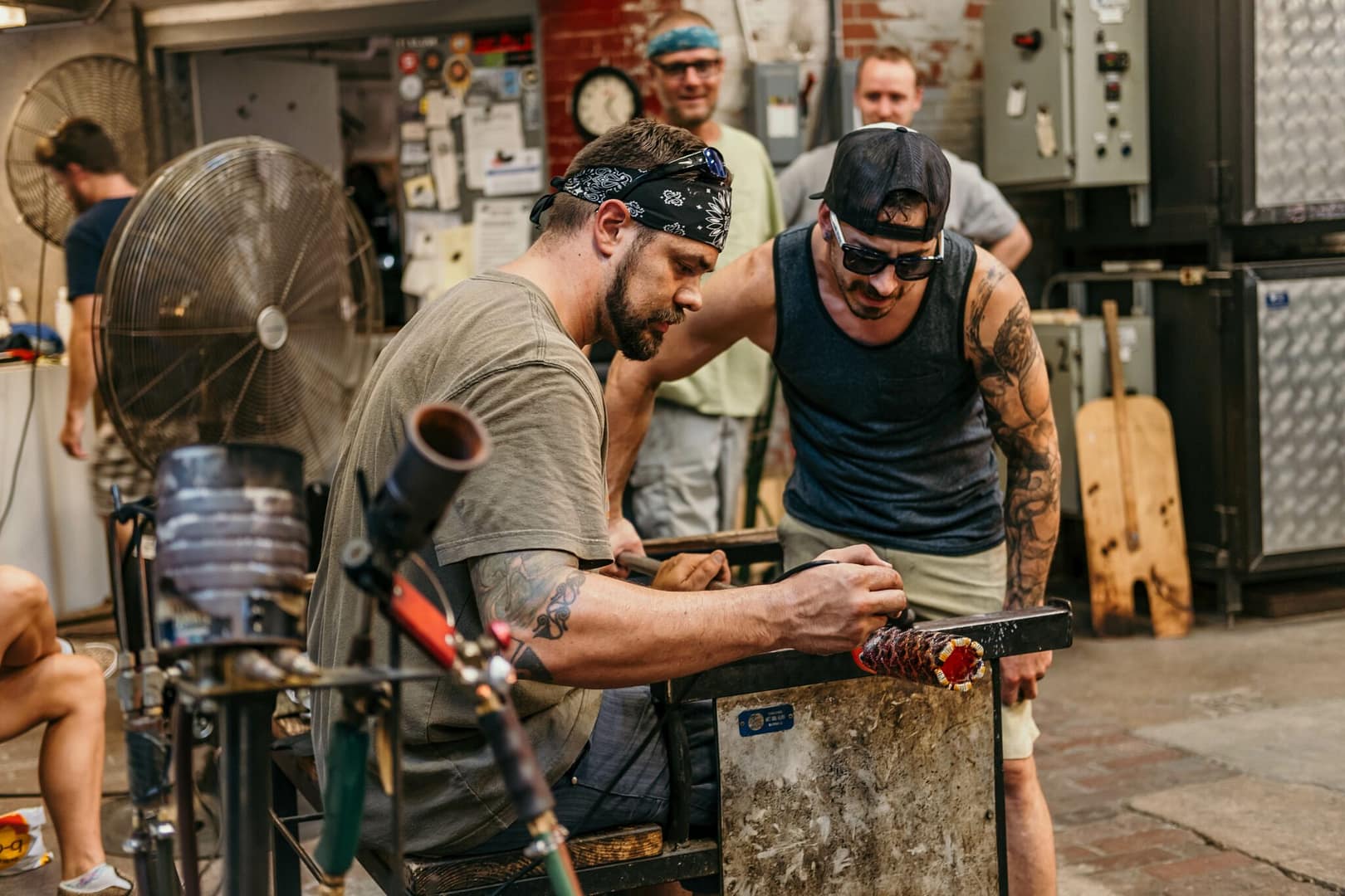 Dan Alexander and a studio assistant looking closely at a glass piece Dan is grinding, while two people observe in the background.