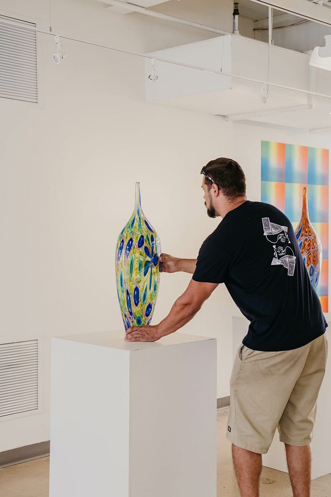 Dan Alexander, placing a tall, narrow piece of glass artwork on a white pedestal. He is turned away from the camera. The artwork is translucent blue and green.